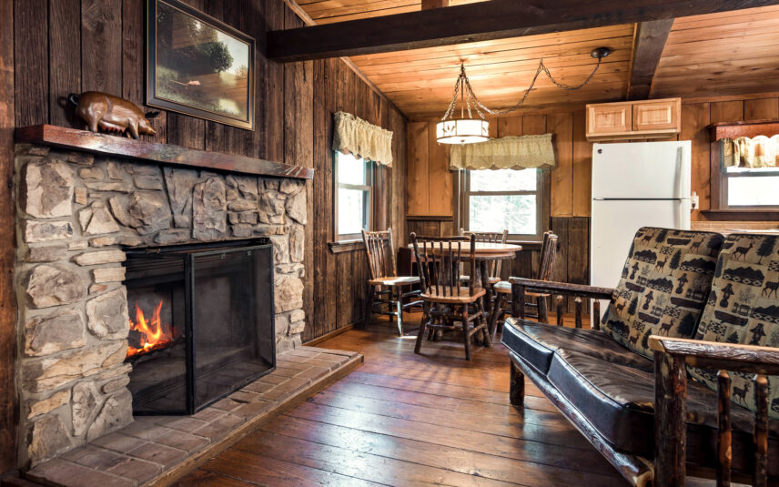 Cozy living room in the Liggett Cabin at Gateway Lodge in Cook Forest, Pennsylvania, featuring a stone fireplace with a crackling fire, a rustic log-style sofa with patterned cushions, a round wooden dining table with chairs, and wood-paneled walls and ceiling.