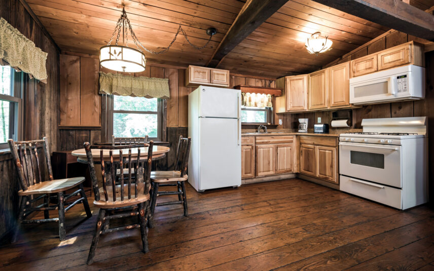 Kitchen and dining area in the Liggett Cabin at Gateway Lodge in Cook Forest, Pennsylvania, featuring light wood cabinets, white appliances (refrigerator, microwave, stove), a round wooden dining table with four chairs, and wood-paneled walls and ceiling.