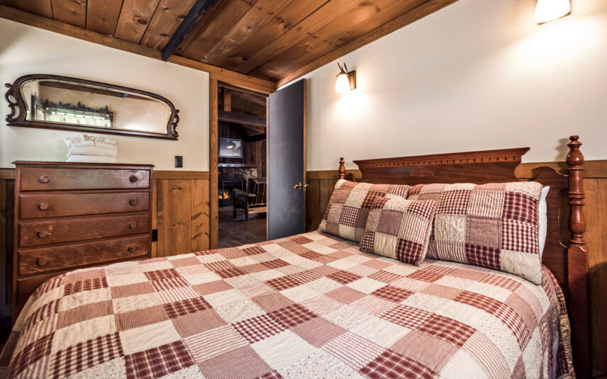 Cozy bedroom with a queen bed featuring a patchwork quilt in the Liggett Cabin at Gateway Lodge in Cook Forest, Pennsylvania. A wooden dresser with a mirror and a glimpse into the living room with a fireplace are visible.