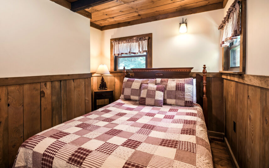 Cozy bedroom in the Liggett Cabin at Gateway Lodge in Cook Forest, Pennsylvania, featuring a queen bed with a patchwork quilt, wooden walls, a wooden nightstand with a lamp, and two windows with valances.