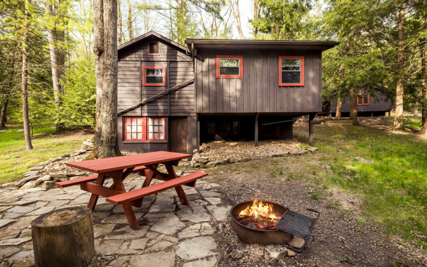 Exterior view of the Liggett Cabin at Gateway Lodge in Cook Forest, Pennsylvania, featuring a dark siding, red window trim, a red picnic table, and a fire pit with a crackling fire in a wooded setting.