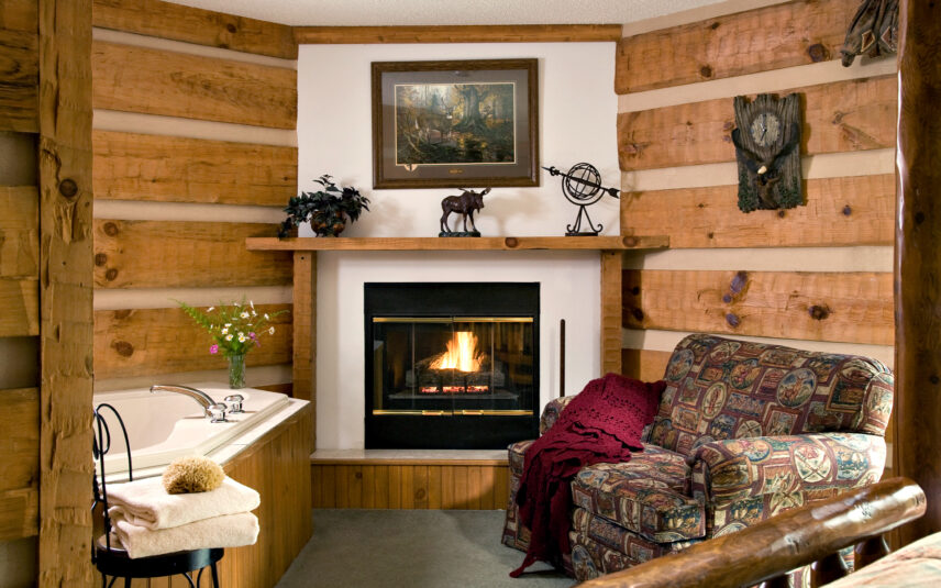 Luxurious Fireside Suite at Gateway Lodge in Cook Forest, Pennsylvania, featuring a cozy gas fireplace with a wooden mantel, a corner jetted tub, a comfortable patterned loveseat with a throw blanket, and rustic hand-hewn beams creating a warm, romantic ambiance.