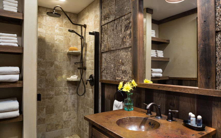 Luxurious bathroom in a Gallery Room at Gateway Lodge in Cook Forest, Pennsylvania, featuring a unique bark-lined accent wall, a rainfall shower head, a distinctive copper vessel sink on a stone counter, and wood-paneled walls, suggesting a rustic yet elegant lodge aesthetic.
