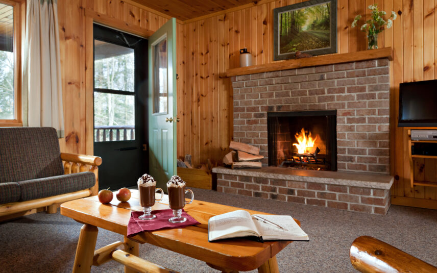 Cozy living room in a Black Bear Cabin at Gateway Lodge in Cook Forest, Pennsylvania, featuring a lit brick fireplace, a wooden coffee table with two glasses of hot chocolate topped with whipped cream, a book, and comfortable seating.