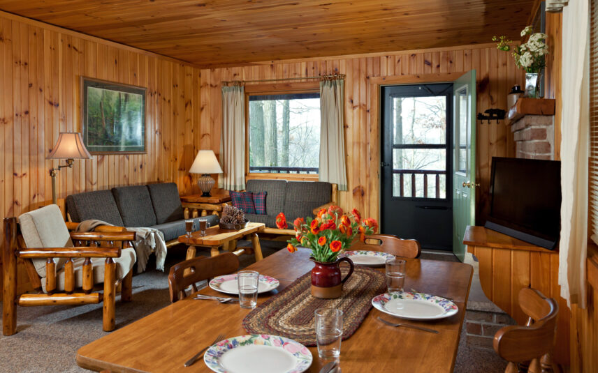 Living and dining area in a Black Bear Cabin at Gateway Lodge in Cook Forest, Pennsylvania, featuring wooden walls and ceiling, two sofas, a wooden dining table set for four, a television, and free wifi access.