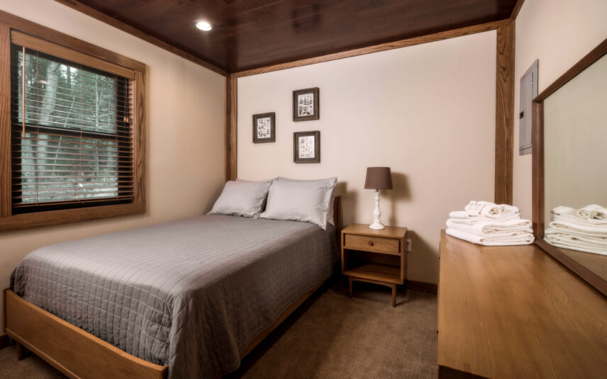 View of the ADA-accessible bedroom in the Cook Cabin at Gateway Lodge, Cook Forest State Park, Pennsylvania, featuring a comfortable bed with accessible clearance, wooden nightstand with lamp, and a window providing natural light, designed with ease of movement in mind.