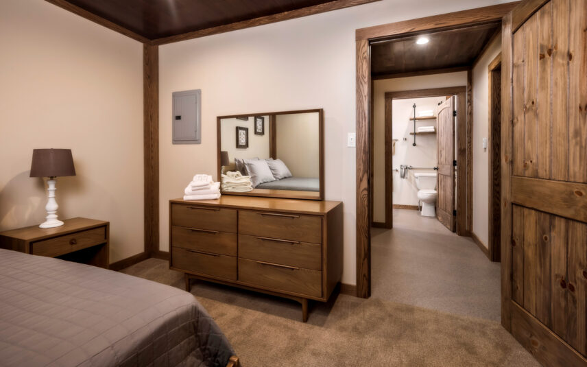 ADA-accessible bedroom of the Cook Cabin at Gateway Lodge in Cook Forest State Park, Pennsylvania, featuring a comfortable bed with neutral bedding, accessible wooden furniture including a dresser and nightstand, and warm lighting, designed for guests with mobility needs.