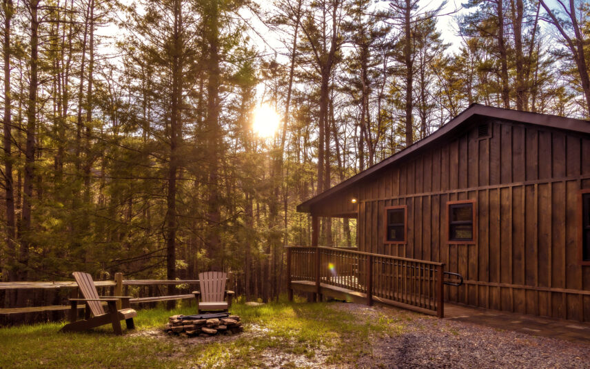 Exterior view of the Cook Cabin, a charming wooden cabin at Gateway Lodge in Cook Forest State Park, Pennsylvania, nestled amongst tall pine and hemlock trees with Adirondack chairs and a stone firepit visible in the yard