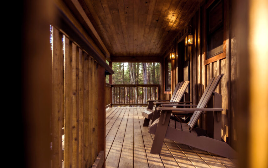 The welcoming wooden porch of the Cook Cabin at Gateway Lodge in Cook Forest State Park, Pennsylvania, featuring two comfortable Adirondack chairs and warm, illuminated outdoor lights.