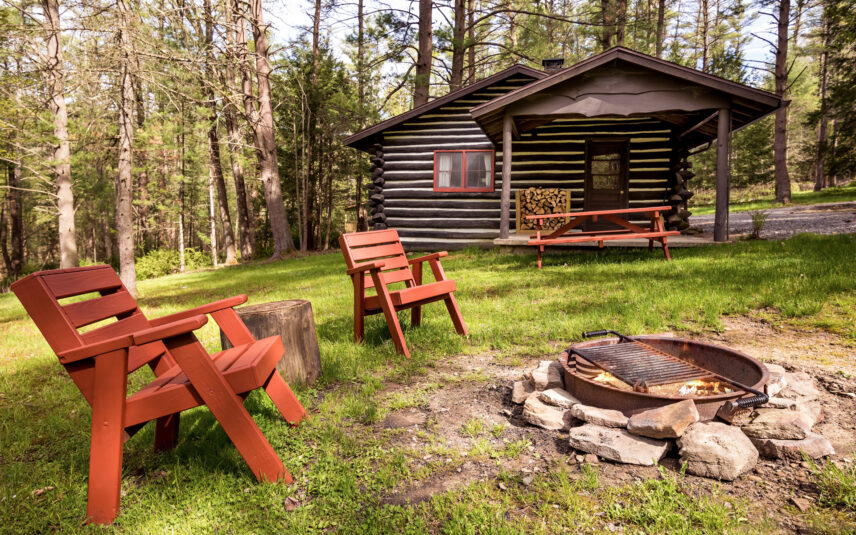 The Hemlock Cabin, a cozy log cabin at Gateway Lodge in Cook Forest State Park, Pennsylvania, featuring two red Adirondack chairs beside a stone firepit with a grill in the foreground.