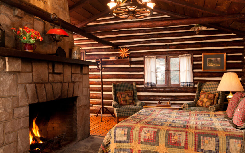 Cozy bedroom with a plaid quilt on the bed in front of a glowing stone fireplace in a log beam Hemlock Cabin at Gateway Lodge, Cook Forest, Pennsylvania. Two plaid armchairs are visible near the bed.
