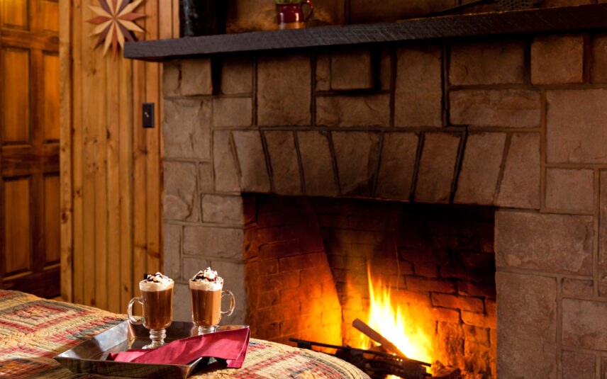 Glowing stone fireplace in a Hemlock Cabin at Gateway Lodge, Cook Forest, Pennsylvania, with a tray holding two mugs of hot chocolate topped with whipped cream on a nearby bed with a colorful quilt.