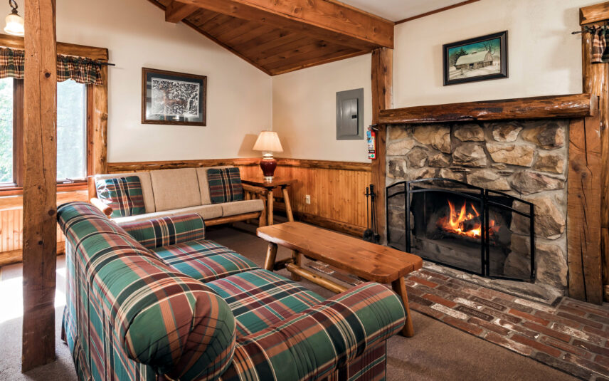 Cozy living room in a Longfellow Cabin at Gateway Lodge in Cook Forest, Pennsylvania, featuring a stone fireplace with a crackling fire, comfortable plaid sofas, wooden beams, and rustic decor.