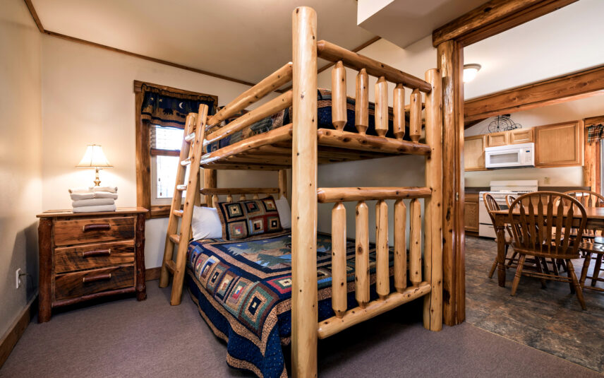 Bedroom in a Longfellow Cabin at Gateway Lodge in Cook Forest, Pennsylvania, featuring a rustic log bunk bed, a wooden nightstand with a lamp, and a window with a dark valance. A glimpse of the cabin's kitchen and dining area is visible.