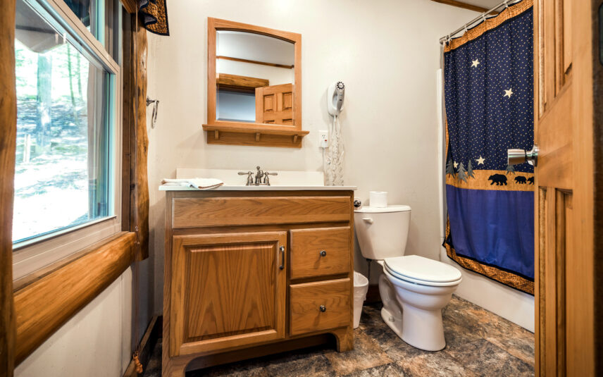 Best Overall Alt Text (Long Tail & SEO-Focused): Bathroom in a Longfellow Cabin at Gateway Lodge in Cook Forest, Pennsylvania, featuring a light wood vanity with a white sink, a wooden-framed mirror, a toilet, and a shower with a blue and gold bear and star-themed curtain.