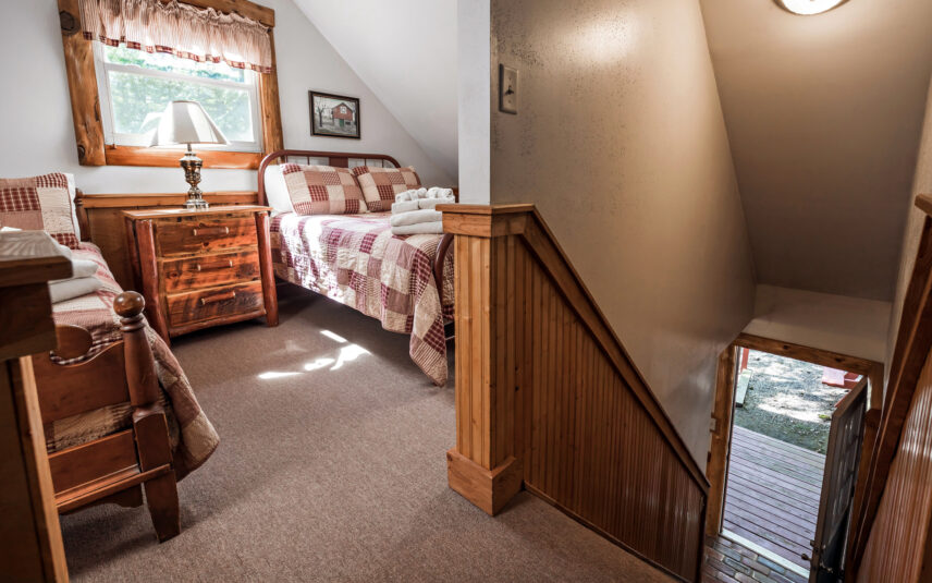 View from the top of the stairs into the loft bedroom of a Longfellow Cabin at Gateway Lodge in Cook Forest, Pennsylvania, showing two beds with patchwork quilts, a wooden nightstand with a lamp, and a window with a valance.