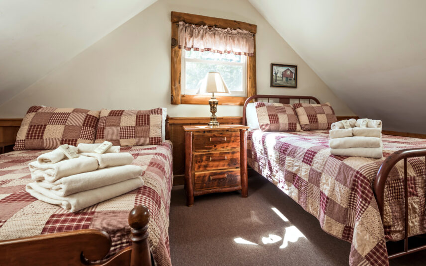 Upstairs loft bedroom in a Longfellow Cabin at Gateway Lodge in Cook Forest, Pennsylvania, featuring two cozy beds with patchwork quilts, a wooden nightstand with a lamp, and a window with a valance.