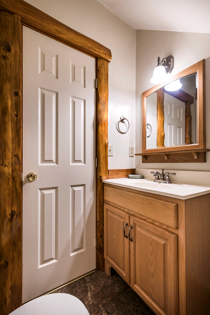 Half bathroom in a Longfellow Cabin at Gateway Lodge in Cook Forest, Pennsylvania, featuring a light wood vanity with a white sink, a wooden-framed mirror, and rustic cedar trim around the door.