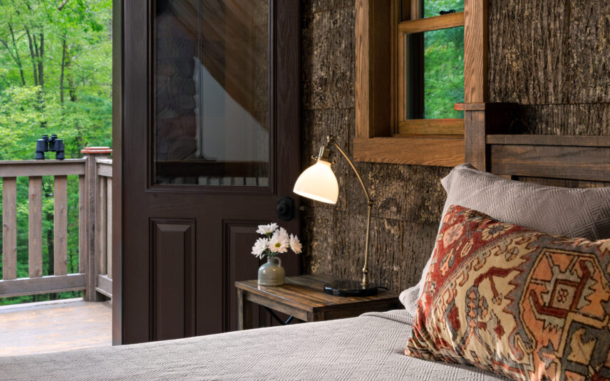Luxury cabin bedroom at Gateway Lodge in Cook Forest, Pennsylvania, featuring a comfortable queen bed, a side table with a lamp and a vase of white daisies, and a small balcony in the background with black binoculars resting on the railing, overlooking the forest.