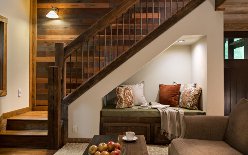 Cozy reading nook with pillows, books, and flowers at Gateway Lodge in Cook Forest, Pennsylvania. Features a built-in bench under the stairs with decorative pillows, a throw blanket, and a glimpse of the rustic cabin interior.