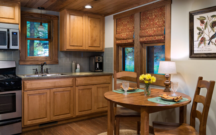 Luxury cabin kitchen at Gateway Lodge in Cook Forest State Park, Pennsylvania. Features modern stainless steel appliances, wooden cabinets, sink, faucet, window with forest view, and a round wooden dining table set for two with yellow flowers. Perfect for a cozy and comfortable getaway in the Pennsylvania Wilds.