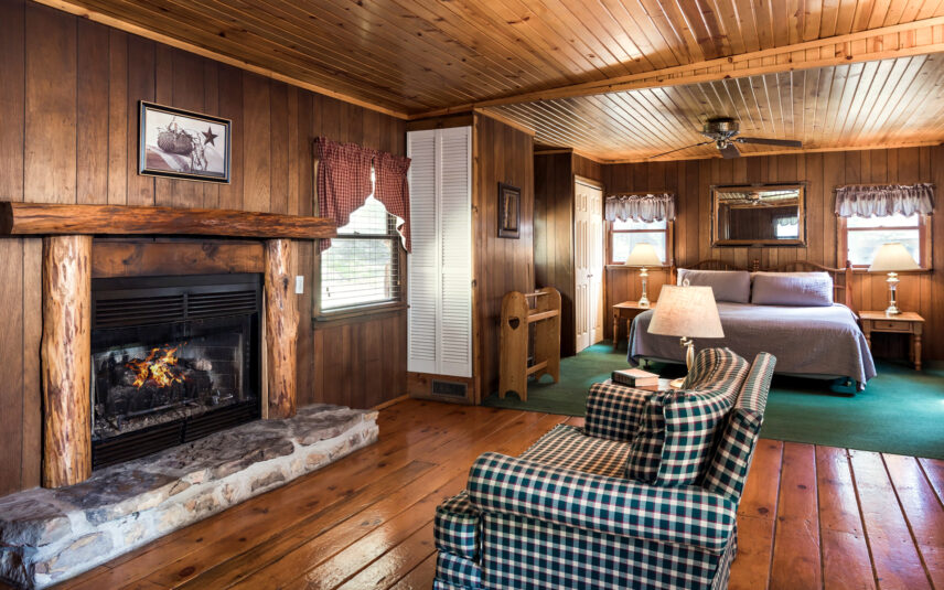 Cozy interior of the Red Eft Cabin at Gateway Lodge in Cook Forest, Pennsylvania, featuring a stone fireplace with a lit gas fire, a plaid armchair, wood-paneled walls and ceiling, and a glimpse into the bedroom with a queen bed.