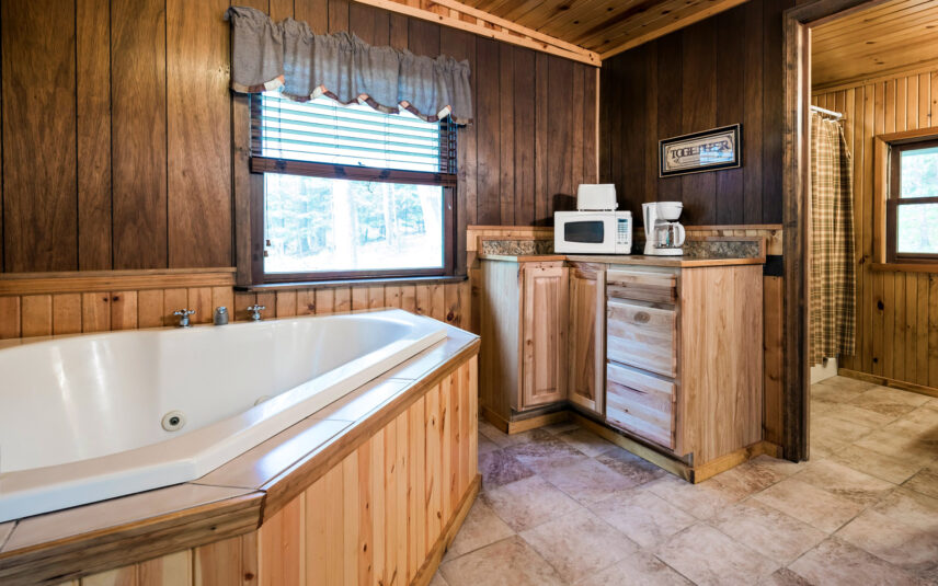 Bathroom with a white jetted tub in the Red Eft Cabin at Gateway Lodge in Cook Forest, Pennsylvania, featuring wood-paneled walls, a window with blinds and a valance, and a glimpse into the shower area.
