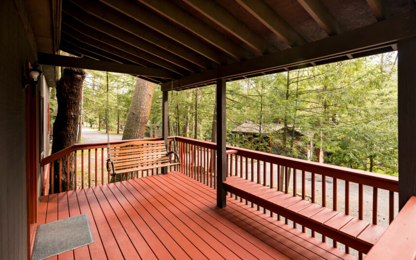 Covered red porch of the Red Eft Cabin at Gateway Lodge in Cook Forest, Pennsylvania, featuring a wooden porch swing, a built-in bench, and a view of the surrounding wooded area.