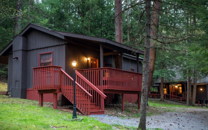 ed Eft Cabin at Gateway Lodge in Cook Forest, Pennsylvania, glowing warmly at night with a red porch and stairs illuminated by outdoor lights, nestled among tall trees.