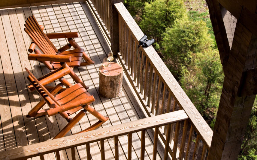 Private suite balcony at Gateway Lodge in Cook Forest, Pennsylvania, featuring two rustic wooden chairs next to a wood stump table holding two wine glasses and black binoculars, offering a peaceful outdoor retreat.