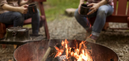 closeup of campfire with two people conversing in the background holding black mugs