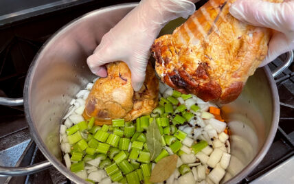 ham being placed in stainless steel pot with chopped celery, carrots and onions