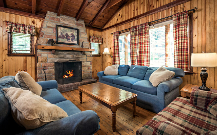 Cozy living room in Tom's Run Cabin at Gateway Lodge in Cook Forest, Pennsylvania, featuring a stone fireplace with a crackling fire, comfortable blue sofas with throw pillows, a wooden coffee table, plaid curtains, and wood-paneled walls and ceiling.