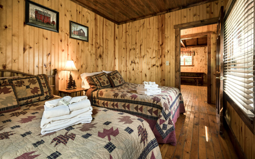 Bedroom with two queen beds featuring patterned quilts in Tom's Run Cabin at Gateway Lodge in Cook Forest, Pennsylvania. Wooden walls and ceiling, a wooden nightstand with a lamp, and a glimpse into another room with a bench are visible.