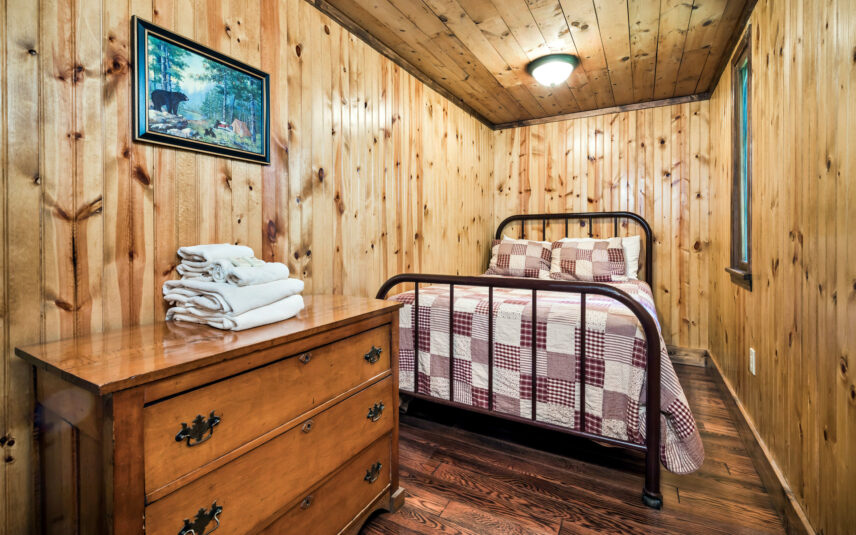 Cozy bedroom in Tom's Run Cabin at Gateway Lodge in Cook Forest, Pennsylvania, featuring a metal frame bed with a patchwork quilt, a wooden dresser with folded towels, and wood-paneled walls and ceiling. A framed picture of a bear hangs on the wall.