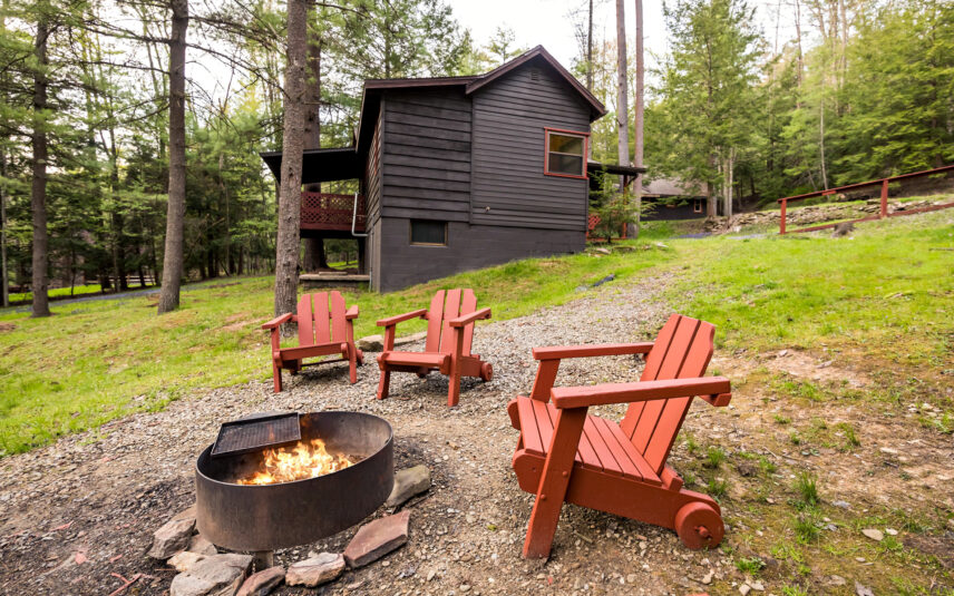 Exterior view of Tom's Run Cabin at Gateway Lodge in Cook Forest, Pennsylvania, featuring a dark siding, red window trim, a red porch, and a stone fire pit with a crackling fire surrounded by red Adirondack chairs in a wooded setting.