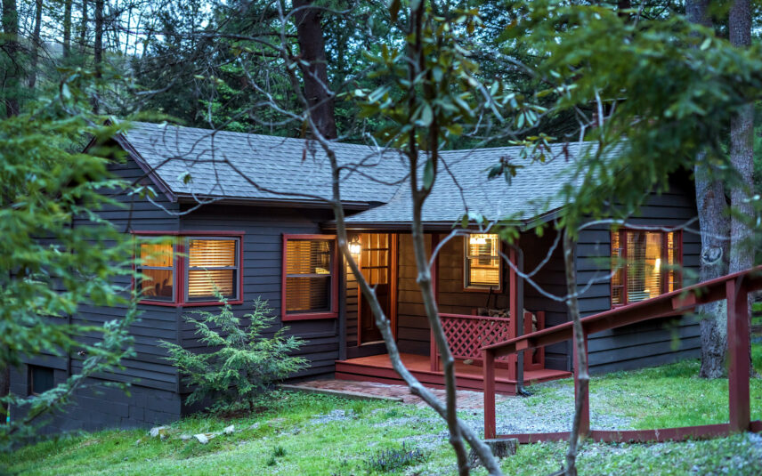 xterior view of Tom's Run Cabin at Gateway Lodge in Cook Forest State Park, Pennsylvania, glowing warmly at dusk with illuminated windows and a red porch, nestled among lush green trees.