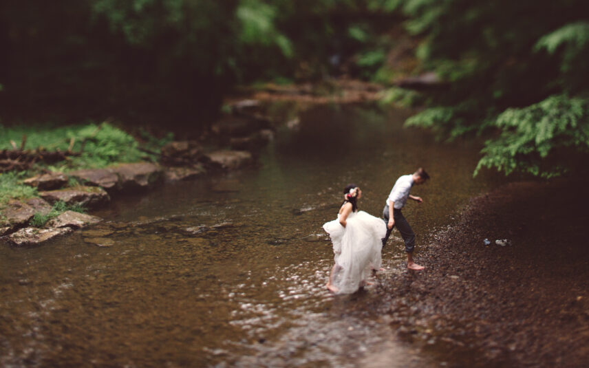 A softly focused photograph capturing a tender moment between newlyweds who may have just celebrated a micro-wedding or elopement. They are wading barefoot through the clear, shallow waters of a creek or river within the romantic setting of Cook Forest State Park in Pennsylvania. The bride, wearing a flowing white wedding dress and colorful flowers in her hair, looks upwards with joy. The groom, in a light-colored collared shirt and darker pants rolled up, gazes down at the water as they step together. The lush green background and gentle ripples emphasize the intimate and peaceful natural ambiance, perfect for small weddings and elopements offered near Gateway Lodge.