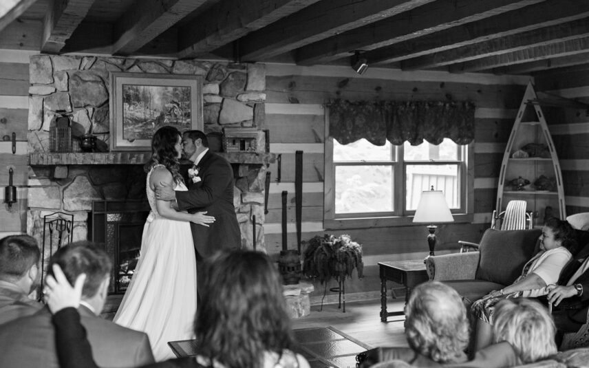 Black and white image of wedding couple kissing in front of Gateway Lodge's great room fireplace with family gathered nearby.