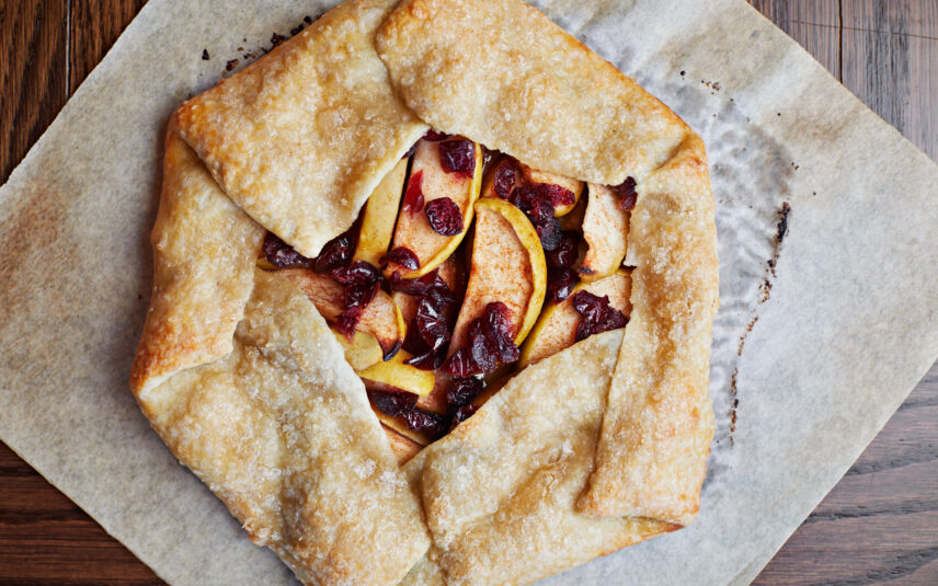 A beautifully presented apple cranberry galette, showcasing slices of baked apples and vibrant red cranberries nestled in a rustic, golden-brown crust. The galette rests on parchment paper on a wooden surface. This homemade dessert, likely from Wild Crafter's Kitchen at Gateway Lodge, highlights the use of fresh, possibly organic and locally sourced ingredients from the PA Wilds region.
