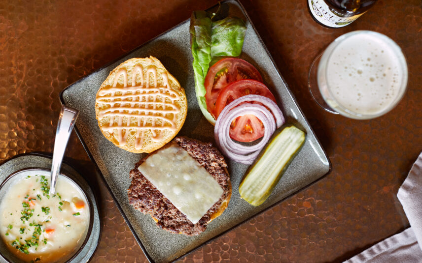 A top-down view of Gateway's Smash Burger, featuring a perfectly seared Clarion Farms ground beef patty topped with melted cheddar cheese, fresh lettuce, ripe tomato slices, crisp dill pickle spears, and thinly sliced red onion, all served on a toasted brioche bun. The meal is accompanied by a bowl of creamy potato soup and a glass of beer. This dish, from Wild Crafter's Kitchen at Gateway Lodge, showcases the use of local ingredients from the PA Wilds.