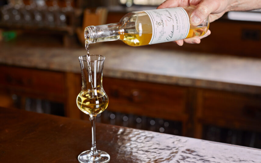 A close-up shot of a hand carefully pouring a golden-hued Sauternes wine from a clear glass bottle into an elegant stemmed glass. The label on the bottle is partially visible and indicates 