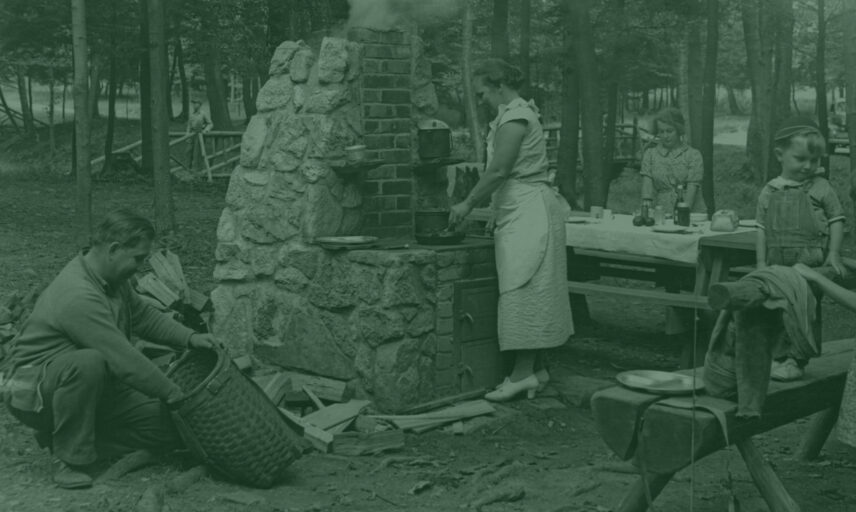 Vintage photograph of a family enjoying an outdoor meal in a wooded setting, possibly within Cook Forest State Park. A woman, dressed in a light-colored dress and apron, is cooking on a unique stone cooktop, with smoke rising from the fire. Her family, including children, are seated at a nearby picnic table, seemingly awaiting their meal. A man is attending to a basket, perhaps containing provisions. The scene evokes a sense of rustic charm and the joys of outdoor dining, reminiscent of a bygone era. This image captures the essence of a family getaway in a natural setting, similar to the experience offered at Gateway Lodge.