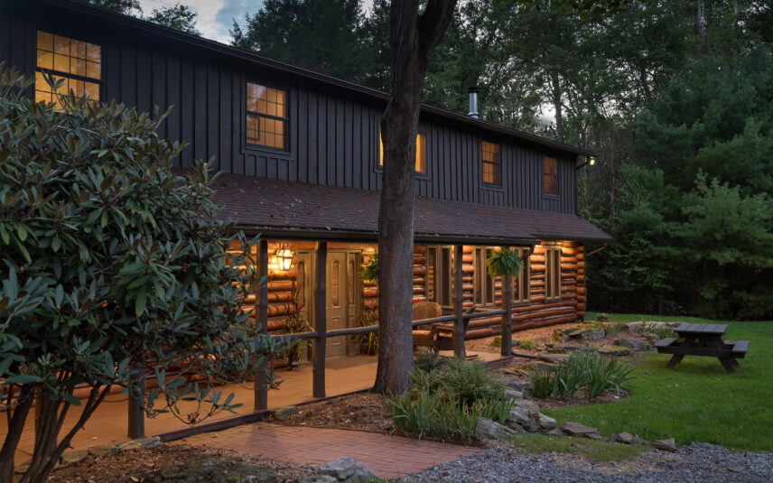 Illuminated wooden log porch entrance to the Forest Meeting Facility and Retreat Center at Gateway Lodge in Cook Forest, Pennsylvania, with a picnic table visible, creating a welcoming ambiance for evening events and group retreats.
