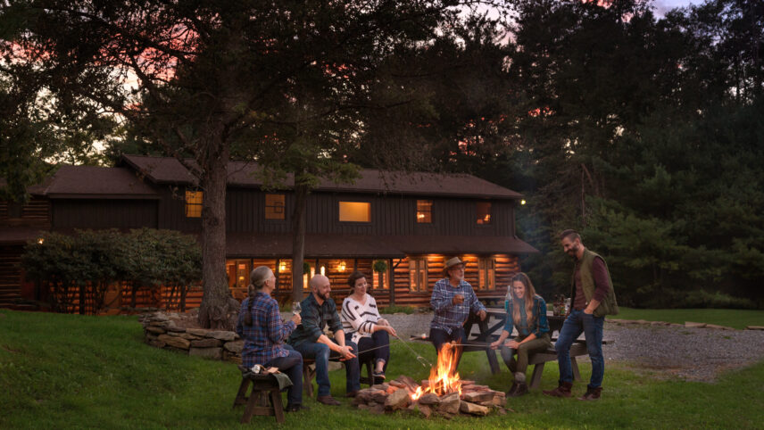 Group of six people roasting s'mores around a campfire in front of a forest meeting facility at Gateway Lodge in Cook Forest, Pennsylvania, with lights illuminating from the facility under beautiful purple evening skies, showcasing group activities and amenities.