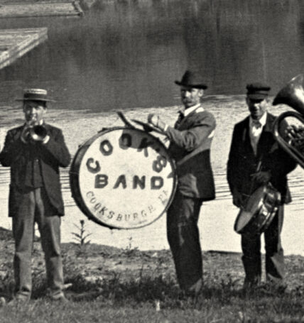 vintage black and white photo of three musicians wearing top hats, playing music near the Clarion River, with the middle man holding a drum labeled 