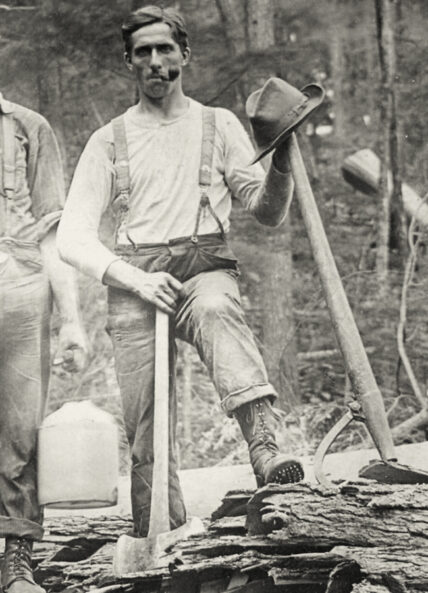 vintage image of a middle-aged man with smoking pipe in his mouth, wearing suspenders and boots, leaning on two axes