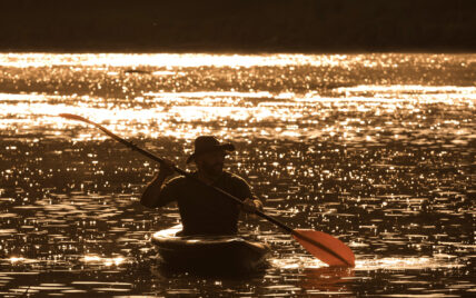 man in a kayak with a sun hat and yellow oars on the Clarion River in the morning, with the sunrise reflecting on the water in the background