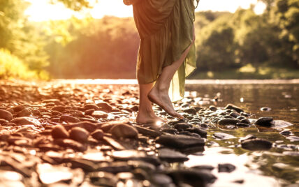 close-up of a barefoot woman in a green dress stepping onto river stones near the Clarion River in Cook Forest State Park on an early morning