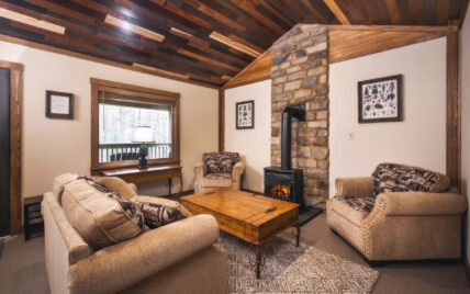 Comfortable and accessible living room in the ADA-compliant Cook Cabin at Gateway Lodge, Cook Forest State Park, Pennsylvania, featuring a cozy sofa, armchairs with space for maneuverability, a wood-burning stove, and a window offering natural light.